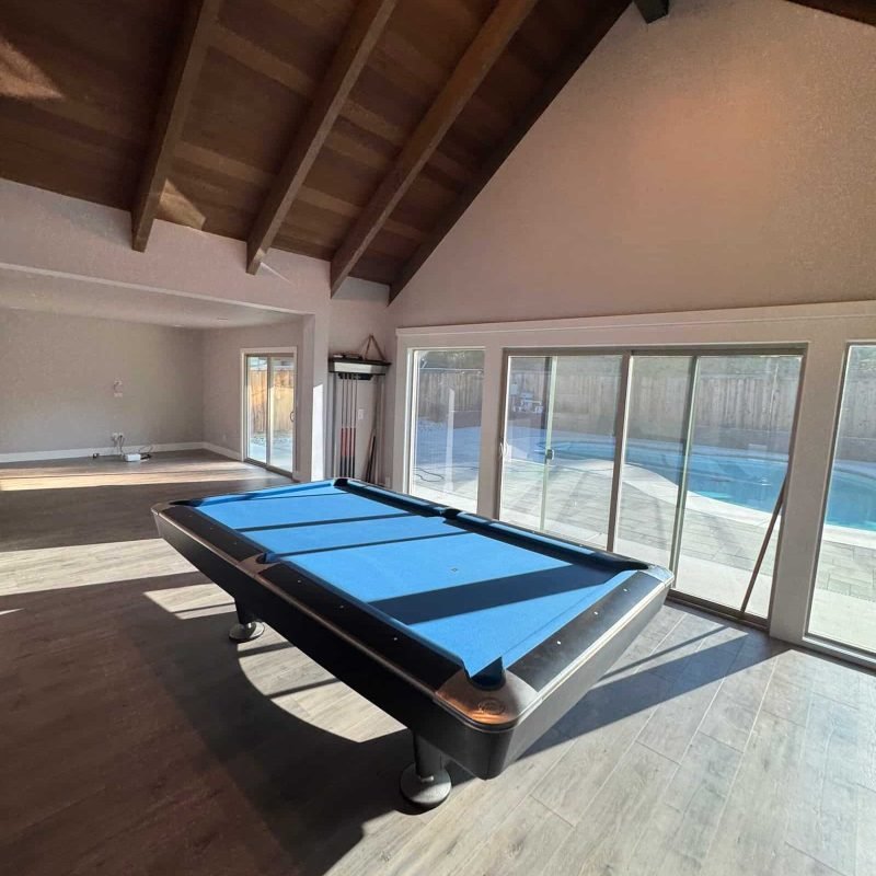 Pool table in a sunlit game room with large windows and vaulted ceiling.